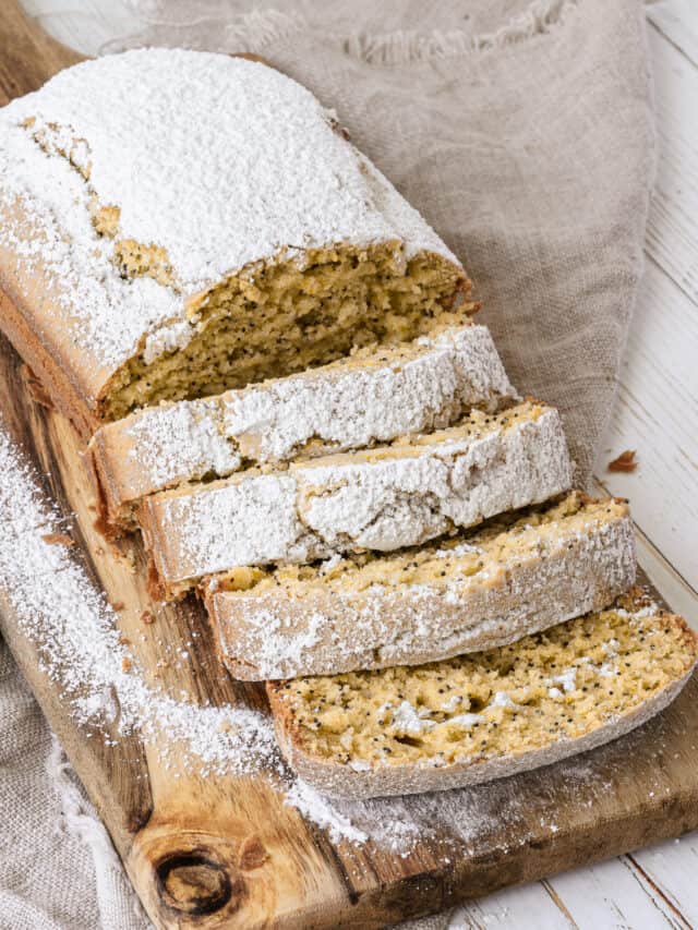 A loaf of sliced lemon poppy seed bread dusted with powdered sugar on a wooden cutting board.
