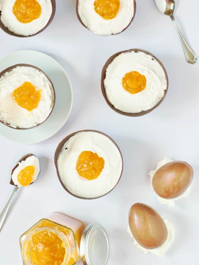 Six bowls of dessert topped with whipped cream and a dollop of yellow jam, chocolate eggs, spoons, and a jar of jam on a white surface.