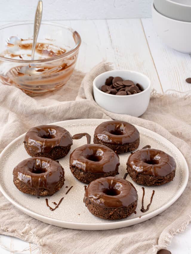 Plate of chocolate donuts with glaze on a wooden table, accompanied by a bowl of chocolate chips and a glass bowl with chocolate spread.