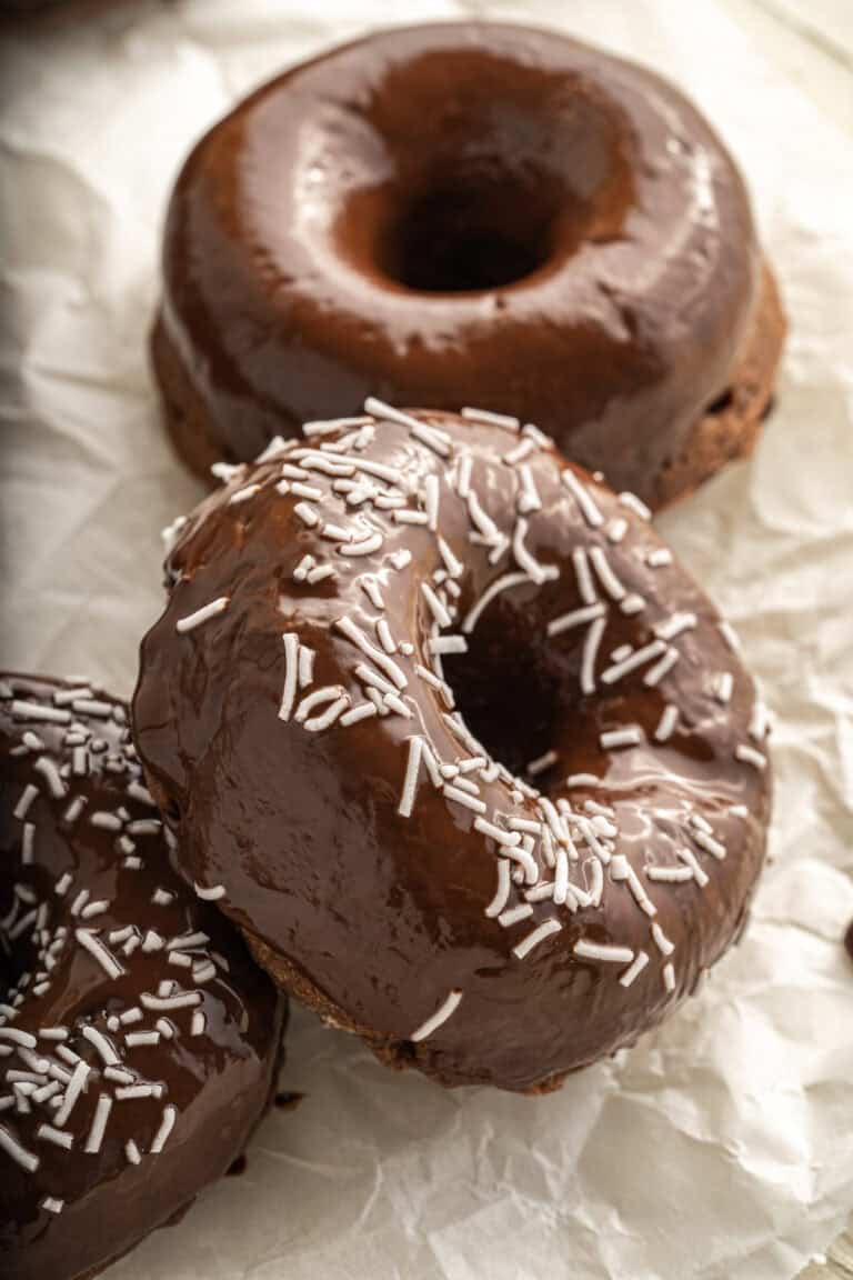 Three vegan chocolate doughnuts, two topped with white sprinkles, arranged on crumpled parchment paper.
