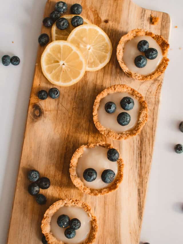 Mini tarts with lemon glaze and blueberries on a wooden board, garnished with fresh blueberries and lemon slices.