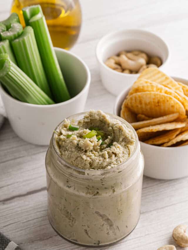 A jar of homemade hummus garnished with green onions, surrounded by celery sticks, crackers, and bowls of nuts and oil.