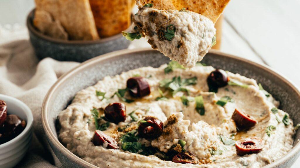 A bowl of eggplant dip with pita chips and olives.