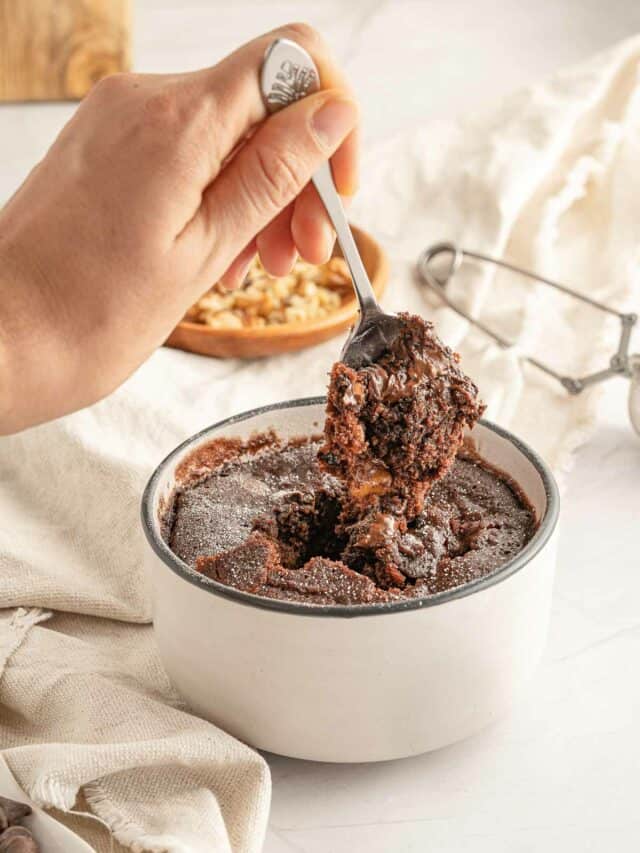 A hand holding a spoon with a chocolate cake in a white bowl.