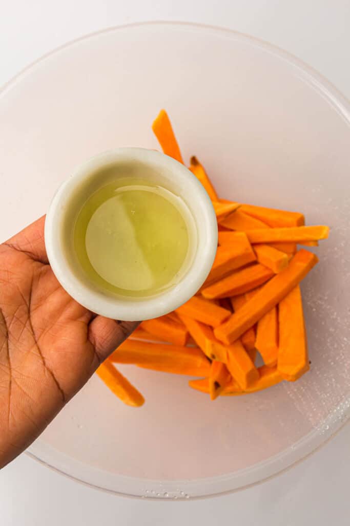 A person holding a cup of oil and slices of sweet potatoes in a bowl.