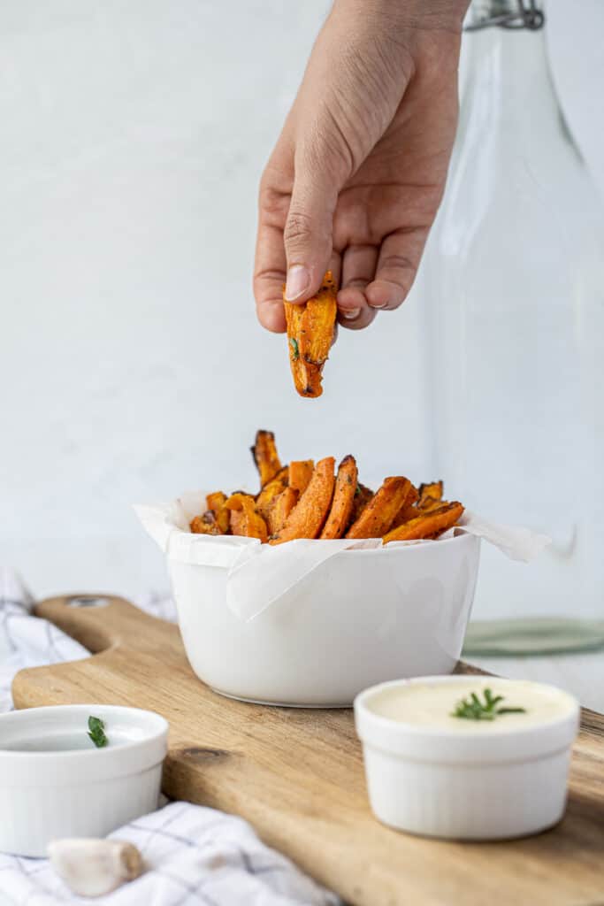 A hand dipping sweet potato fries into a bowl.