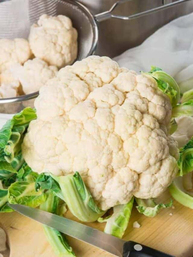 A fresh cauliflower with leaves on a wooden cutting board next to a knife.