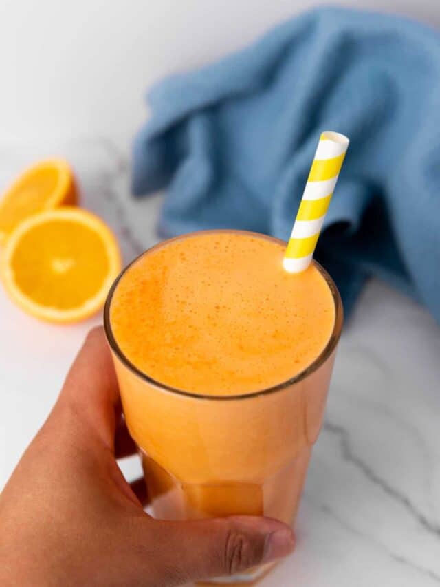 A person holding a glass of orange smoothie with a striped straw.