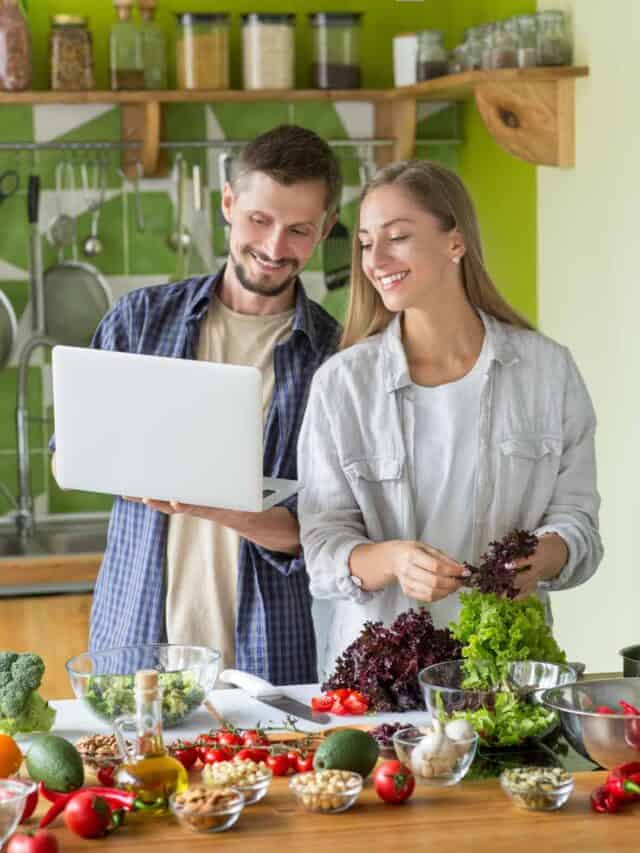 A man and woman standing in front of a kitchen with vegetables and a laptop.