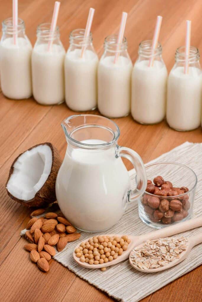 A glass jug of milk with nuts and coconut on a wooden surface.