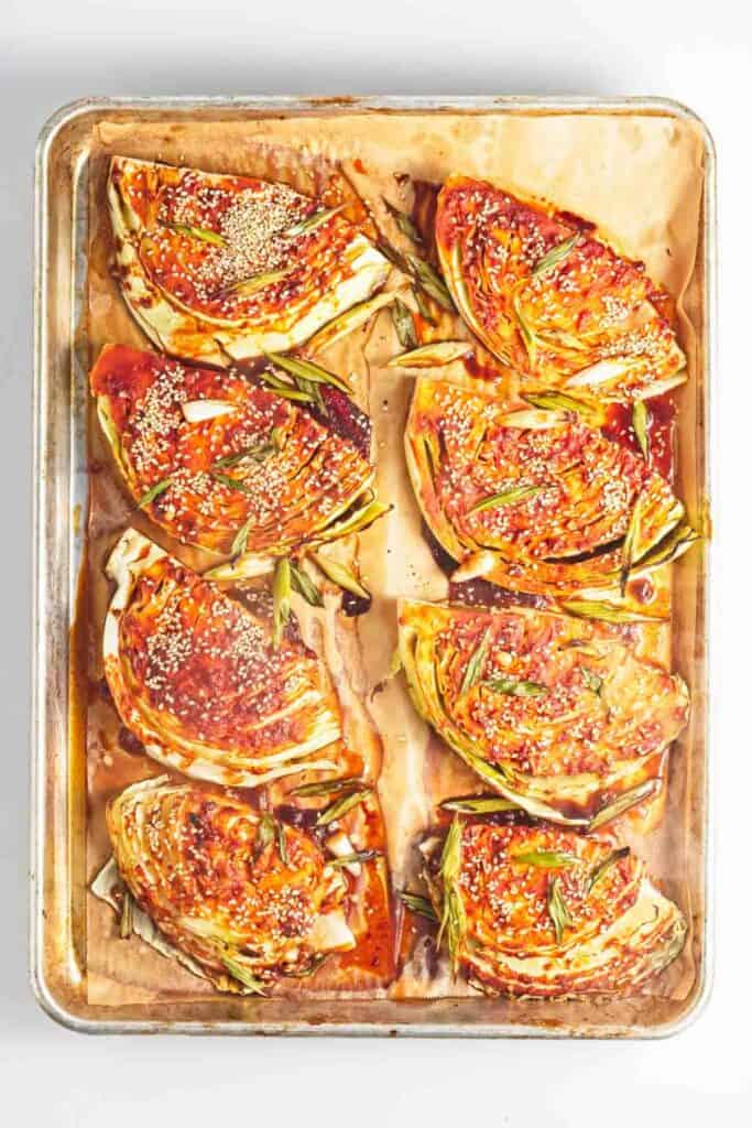 A baking sheet filled with roasted vegetables, including gochujang cabbage steaks, on a white surface.