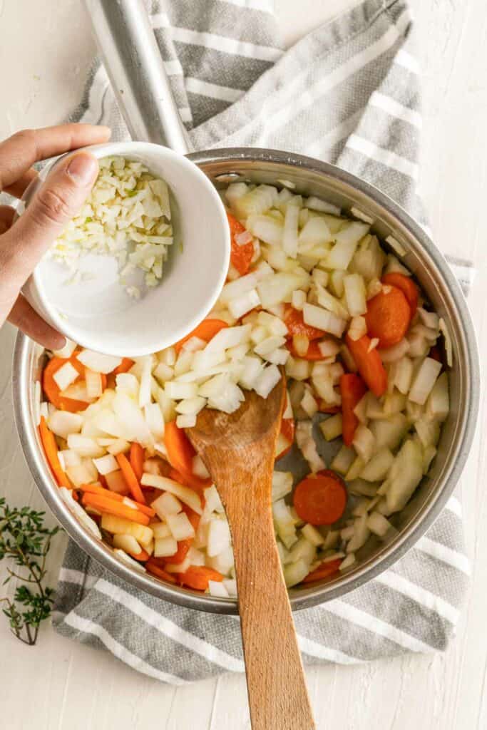A person stirring onions and carrots in a pan with a wooden spoon.