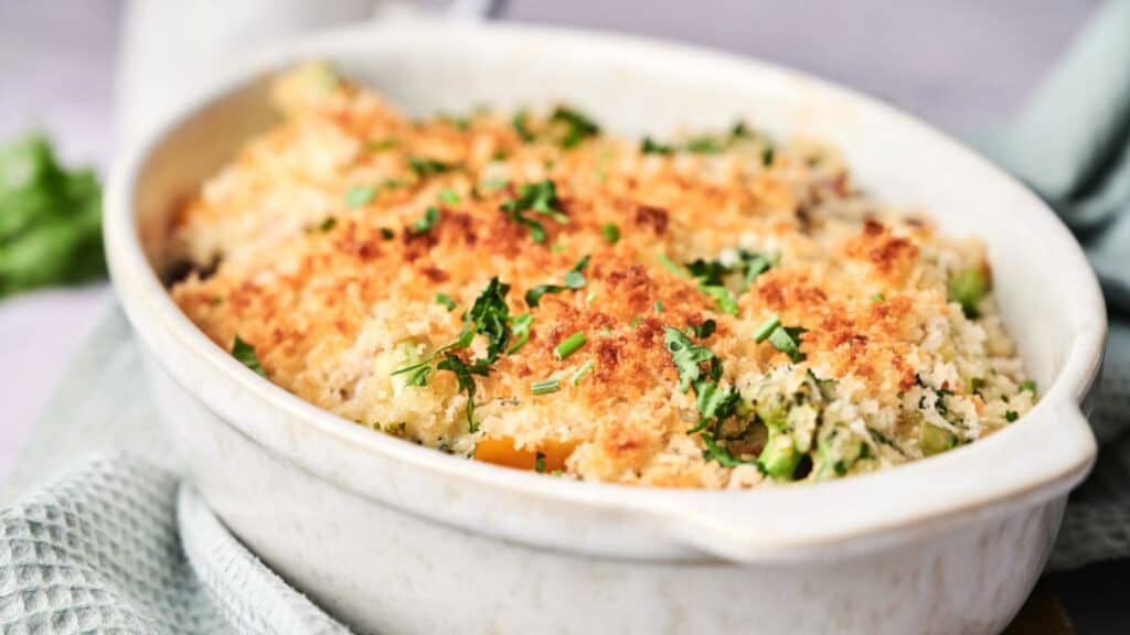 A baked vegetable casserole topped with breadcrumbs and garnished with chopped parsley in a white dish.