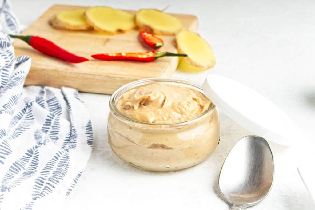 A jar of peanut butter with a spoon beside it, sliced ginger, and red chili peppers on a wooden board, on a white countertop.