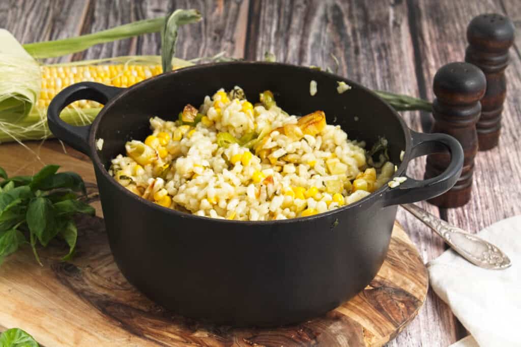 A pot of sweet corn and leek risotto with herbs on a wooden cutting board.