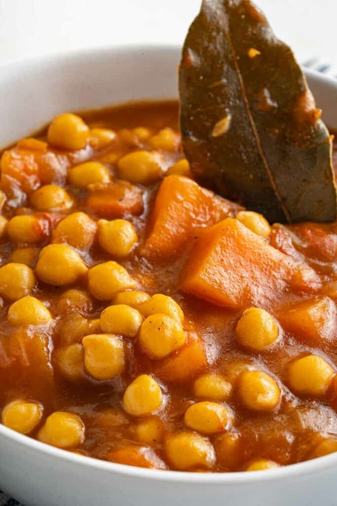 A bowl of stew with chickpeas and a leaf.