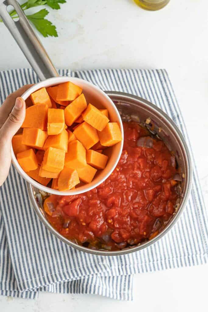 A person pouring tomato sauce into a pan with sweet potatoes.
