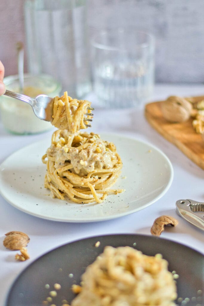 A person is holding a fork to a plate of pasta with nuts.