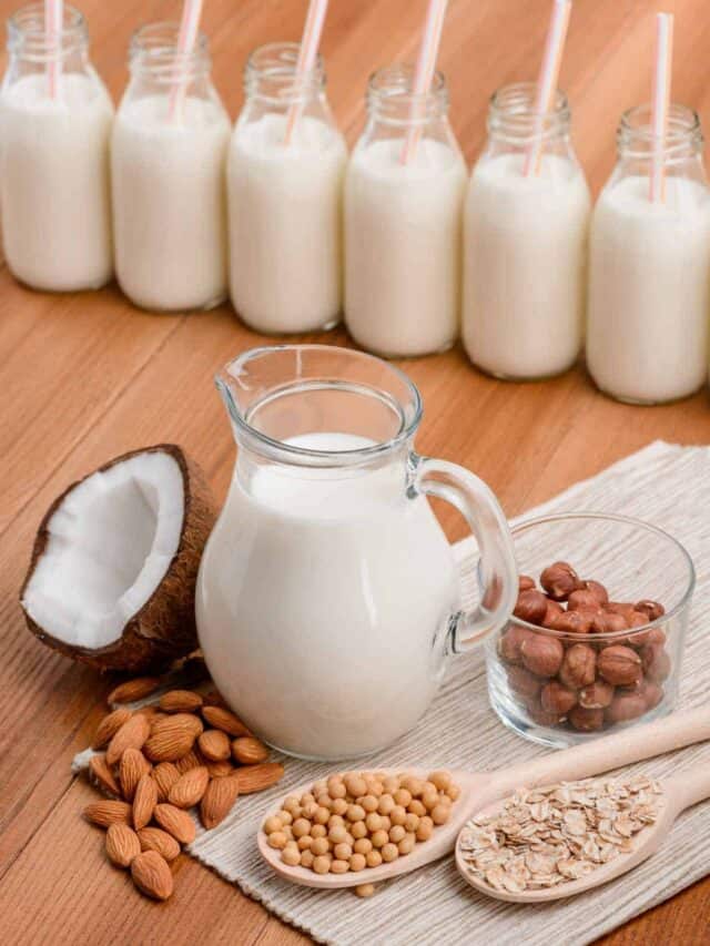 A glass jug of milk with nuts and coconut on a table.