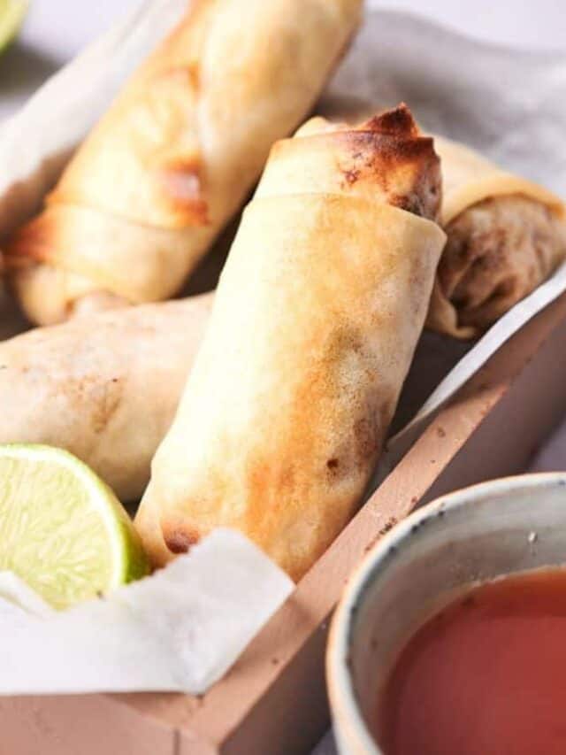 A close-up image of three golden-brown spring rolls in a white tray, served with a slice of lime and a side of dipping sauce.
