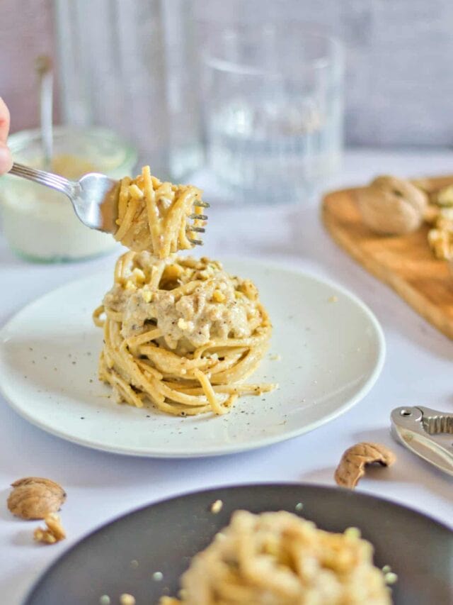 A person is holding a fork to a plate of pasta with nuts on it.