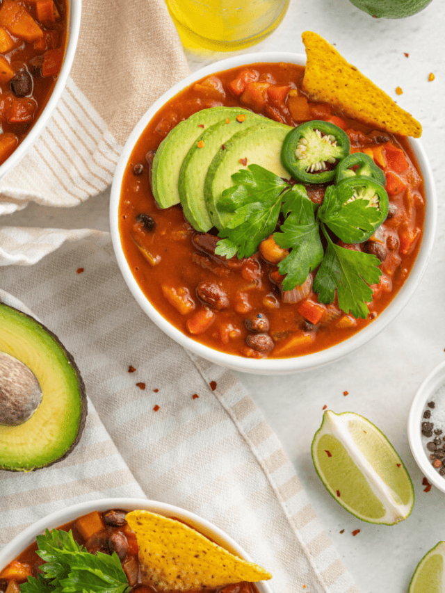 A bowl of chili garnished with avocado slices, cilantro, and a jalapeño, served with tortilla chips and lime wedges on the side.