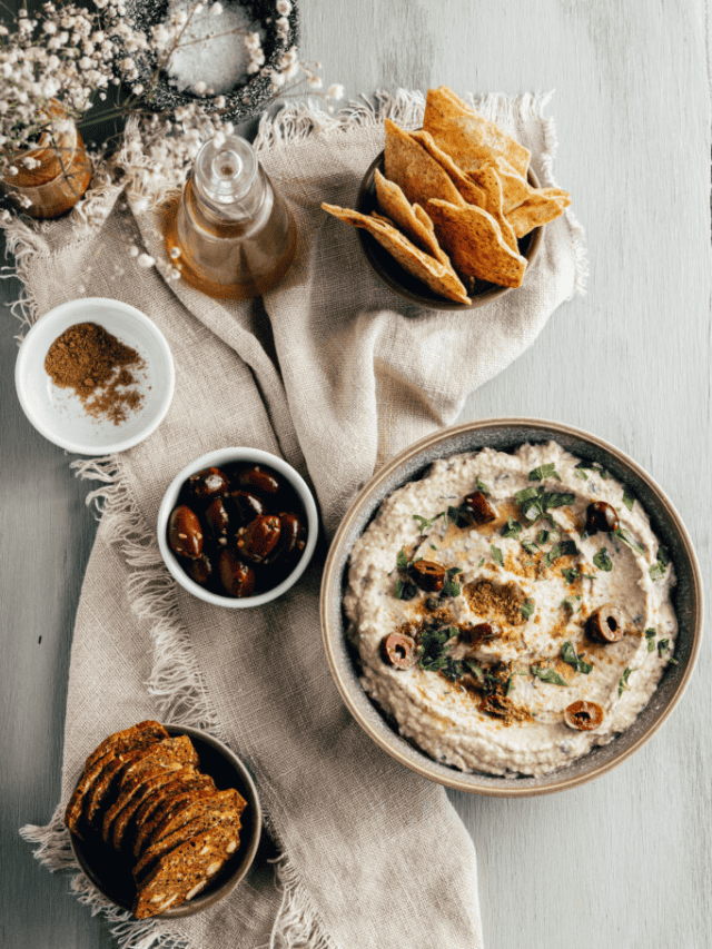 A bowl of hummus garnished with olives and herbs, served with pita chips, slices of bread, and assorted condiments on a rustic table setting.