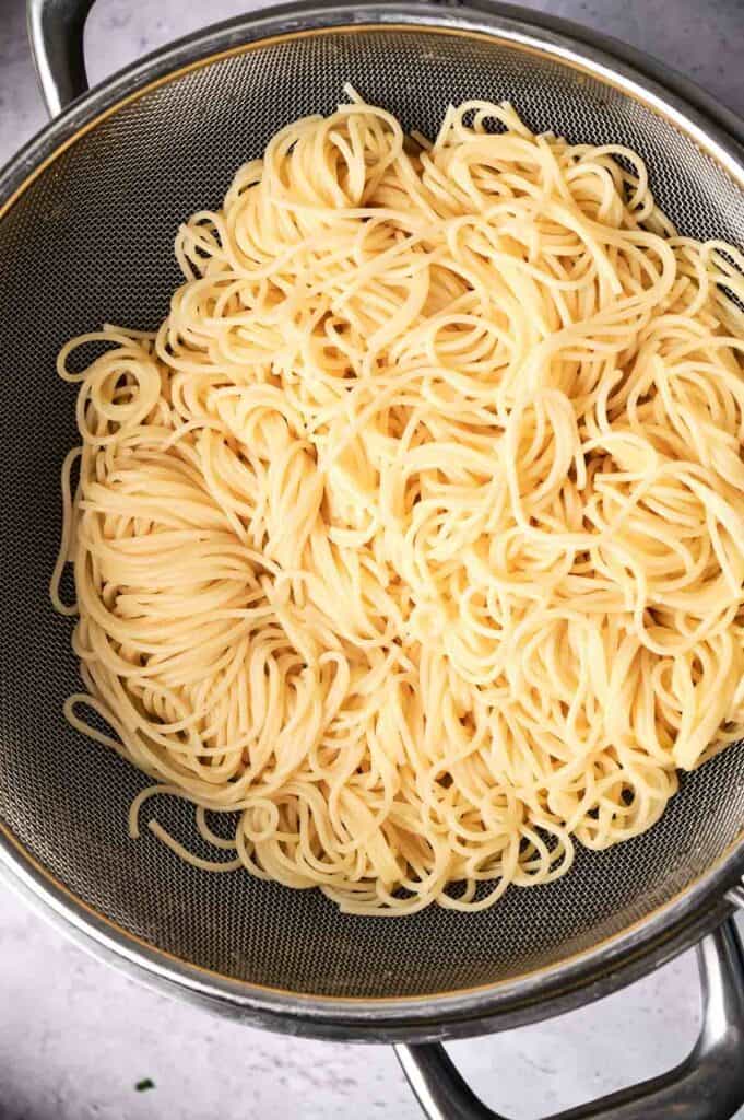 Cooked spaghetti noodles draining in a metal colander placed over a saucepan on a kitchen counter.