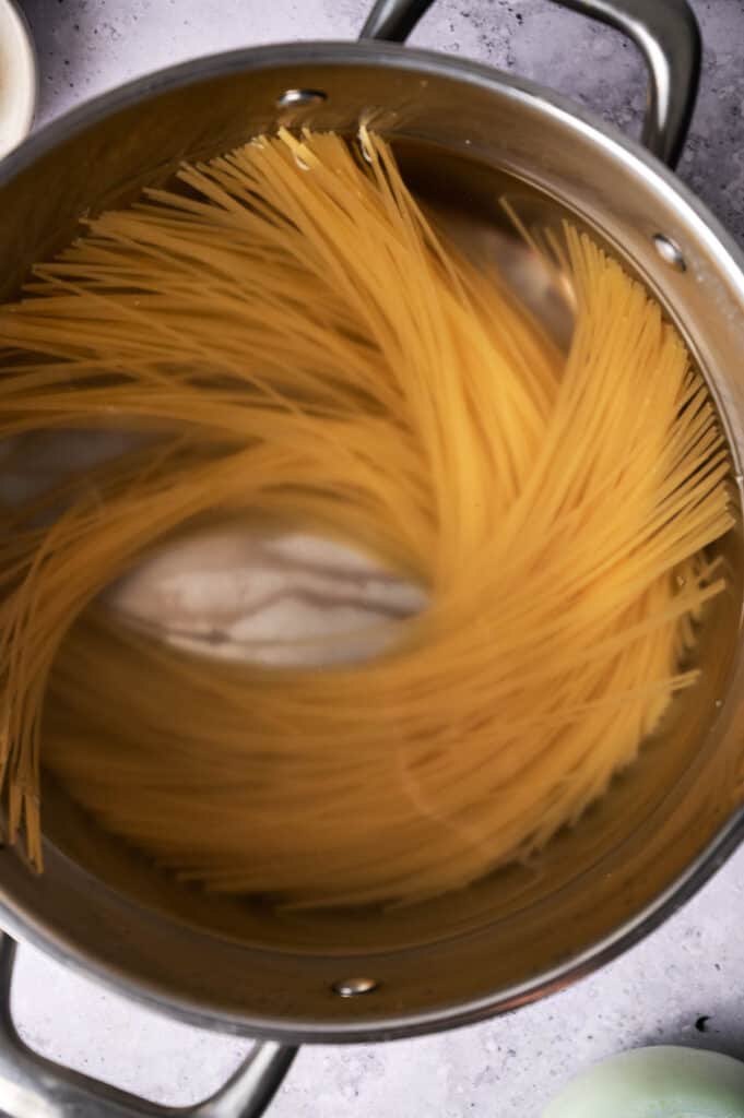 Uncooked spaghetti partially submerged in boiling water in a stainless steel pot.