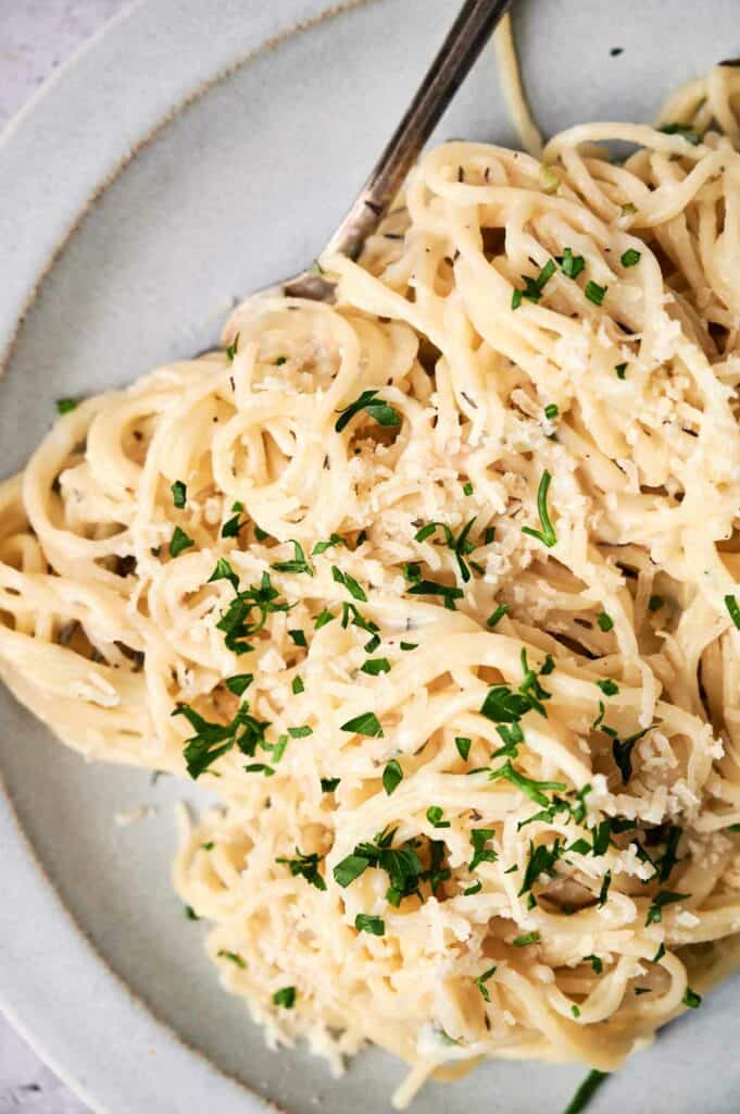 A plate of creamy alfredo pasta topped with grated cheese and garnished with chopped parsley, with a fork on the side.