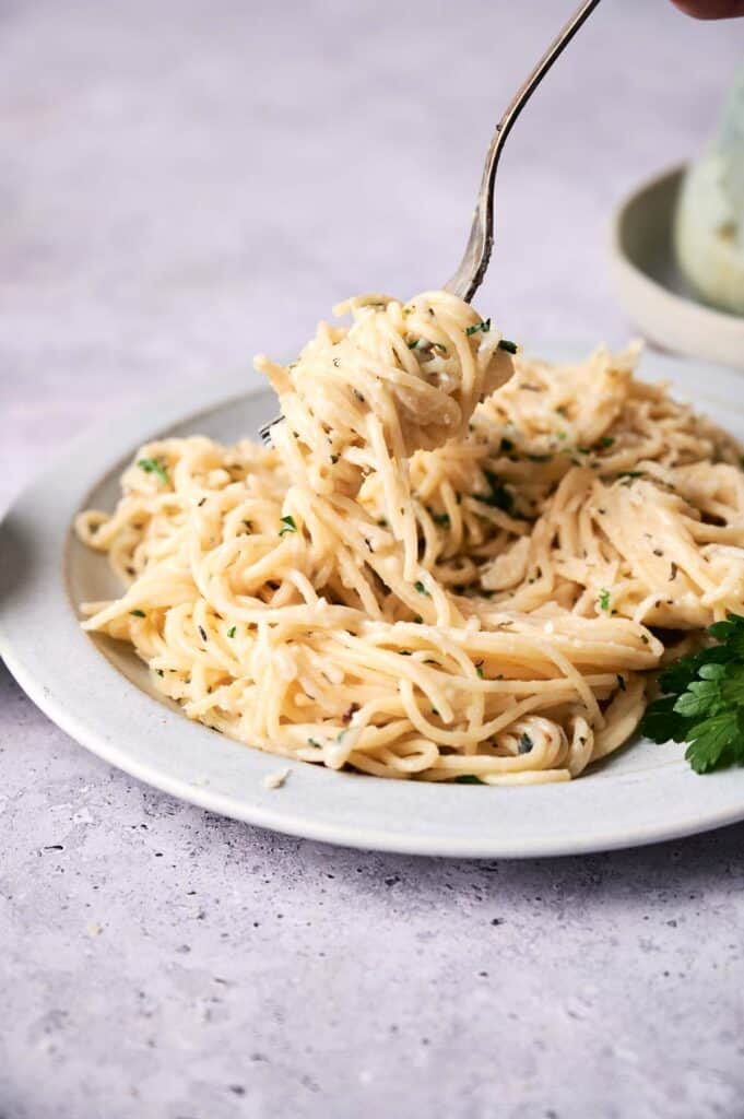 A fork lifting creamy vegan alfredo pasta garnished with herbs from a plate on a light textured surface.