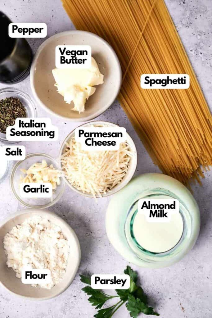 Ingredients for cooking alfredo pasta displayed on a wooden surface including pepper, vegan butter, spaghetti, italian seasoning, parmesan cheese, salt, garlic, flour, parsley, and almond milk, all labeled.