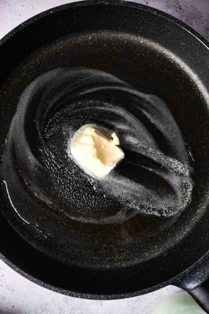 A cube of butter melting in the center of a black non-stick frying pan.