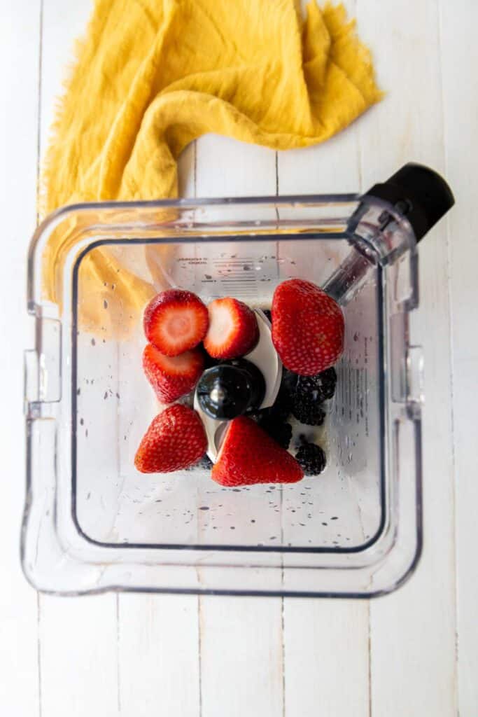 Top view of a blender with strawberries and blackberries on a white wooden surface with a yellow napkin nearby, ready to make a vegan berry smoothie.