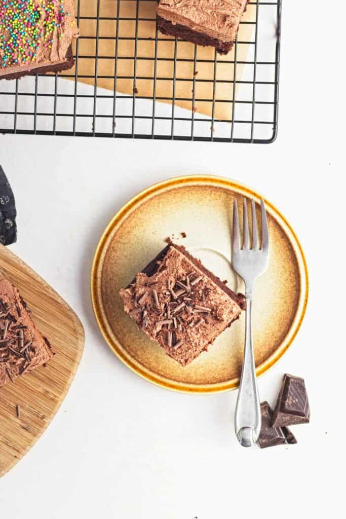 A vegan chocolate cake slice on a ceramic plate with a fork, accompanied by chocolate shavings and other cake pieces in the background.