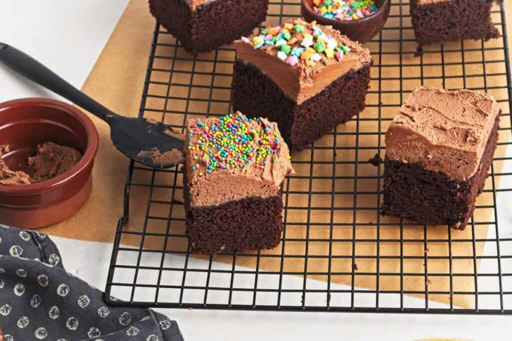 Vegan chocolate cake frosted brownies with sprinkles on a cooling rack, one being served with a spatula.