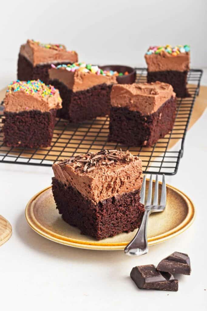 Vegan chocolate cake with sprinkles on a cooling rack, with a single slice served on a plate beside a fork.