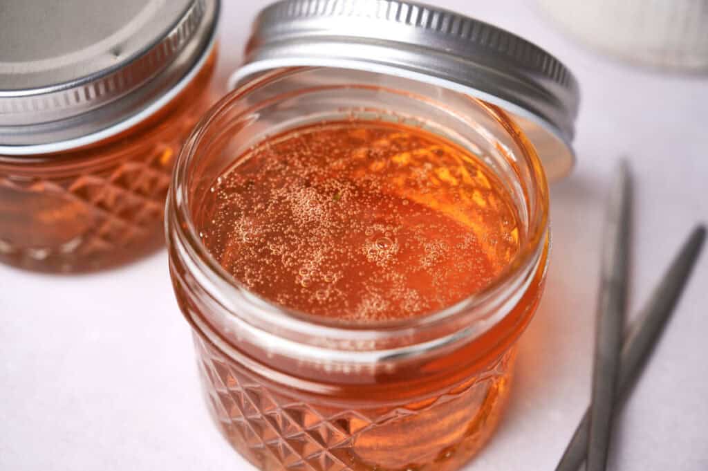 Open jar of vegan honey with bubbles on top, alongside sealed jars and a pair of tongs on a white surface.