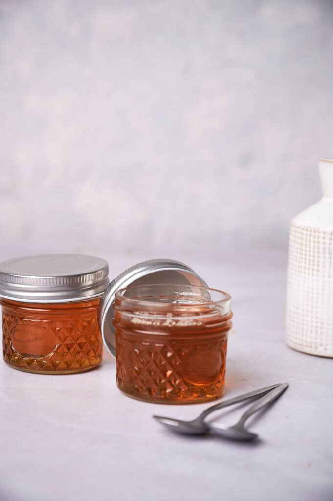 Two jars of vegan honey, one open with a spoon next to it, on a light textured surface against a soft grey background.