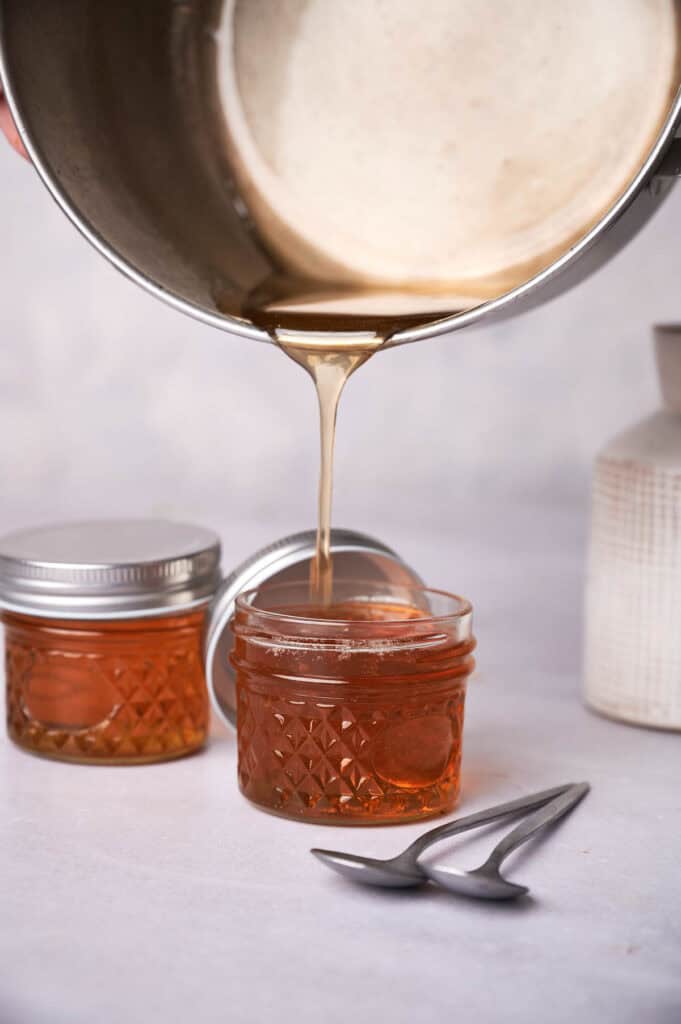 Vegan honey being poured from a metal pot into a small glass jar on a table, with additional jars and a spoon nearby.