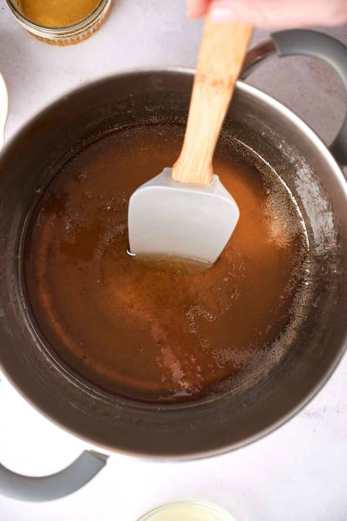 A hand stirs vegan honey with a white spatula in a stainless steel pot on a stove.