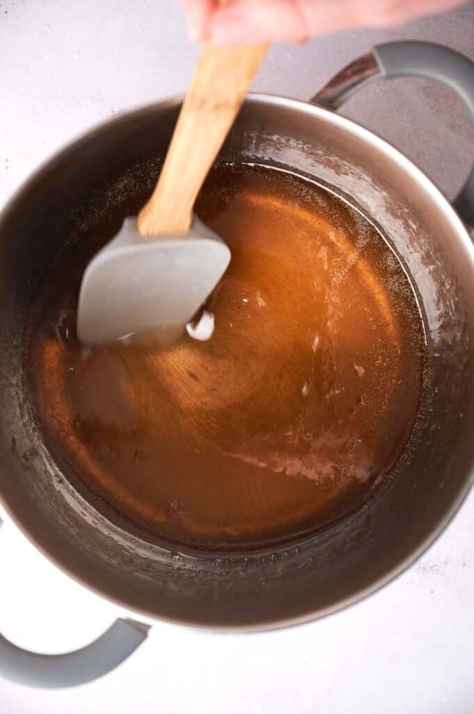 A hand stirring vegan honey with a wooden spoon in a saucepan on a stove.