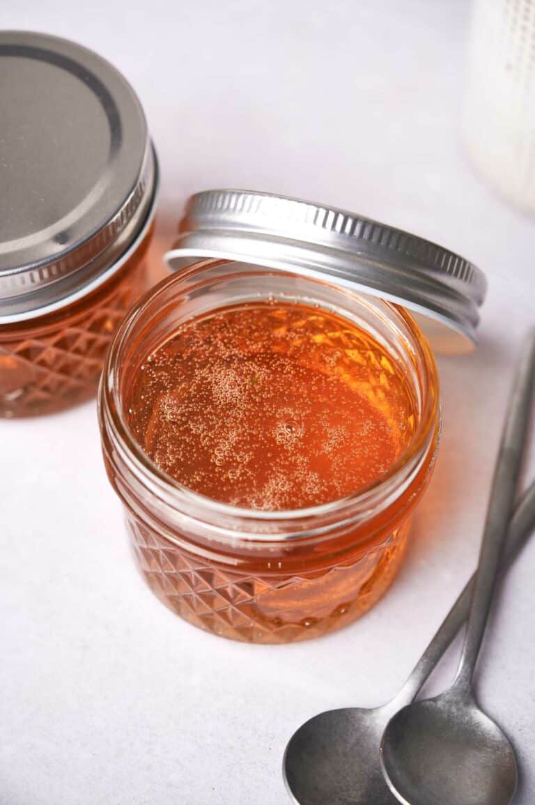 Two jars of vegan honey with metal lids on a white surface, one jar open displaying honey texture, accompanied by two spoons to the side.