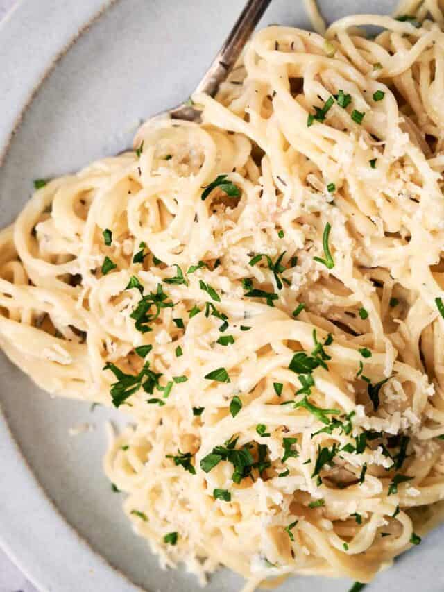 A plate of creamy alfredo pasta topped with grated cheese and garnished with chopped parsley, with a fork on the side.