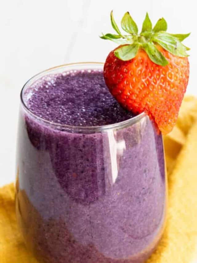 A glass of purple smoothie garnished with a fresh strawberry on the rim, set against a yellow cloth background.