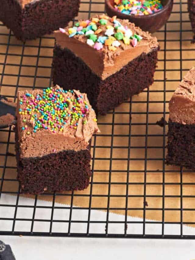 Squares of chocolate frosted cake with colorful sprinkles on a cooling rack, partial view of a bowl with extra sprinkles to the side.