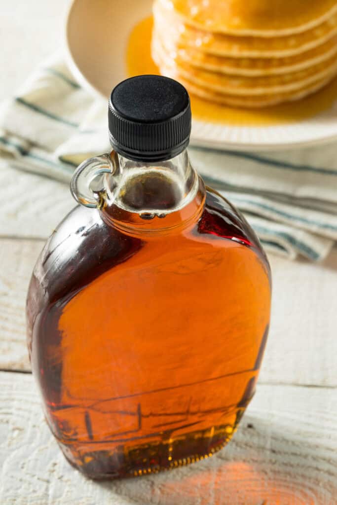 A bottle of amber maple syrup with a black cap in the foreground, with a stack of pancakes in the background on a stripe cloth.