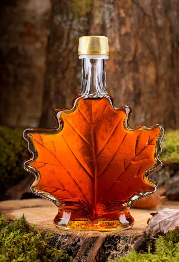 A glass bottle shaped like a maple leaf filled with amber-colored liquid, likely maple syrup, stands on a wooden surface with moss and a tree trunk in the background.