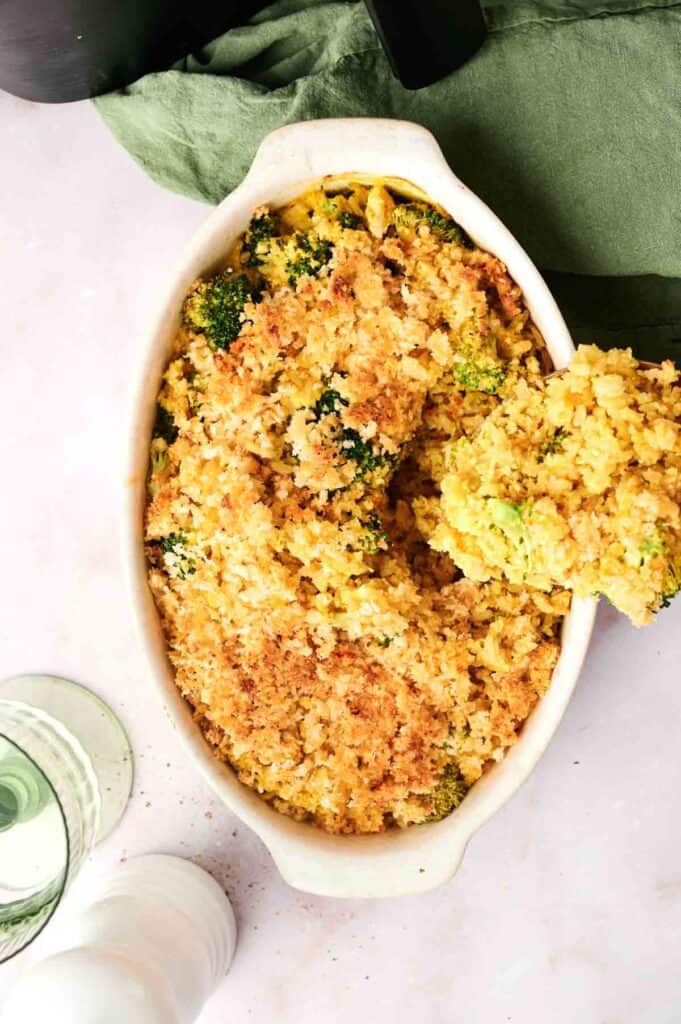 A casserole dish brimming with broccoli cheese casserole, sprinkled with breadcrumbs, sits invitingly alongside a green napkin and a glass of water on a white surface. Consider trying this delightful dish as an air fryer vegan broccoli and rice casserole for a unique twist.