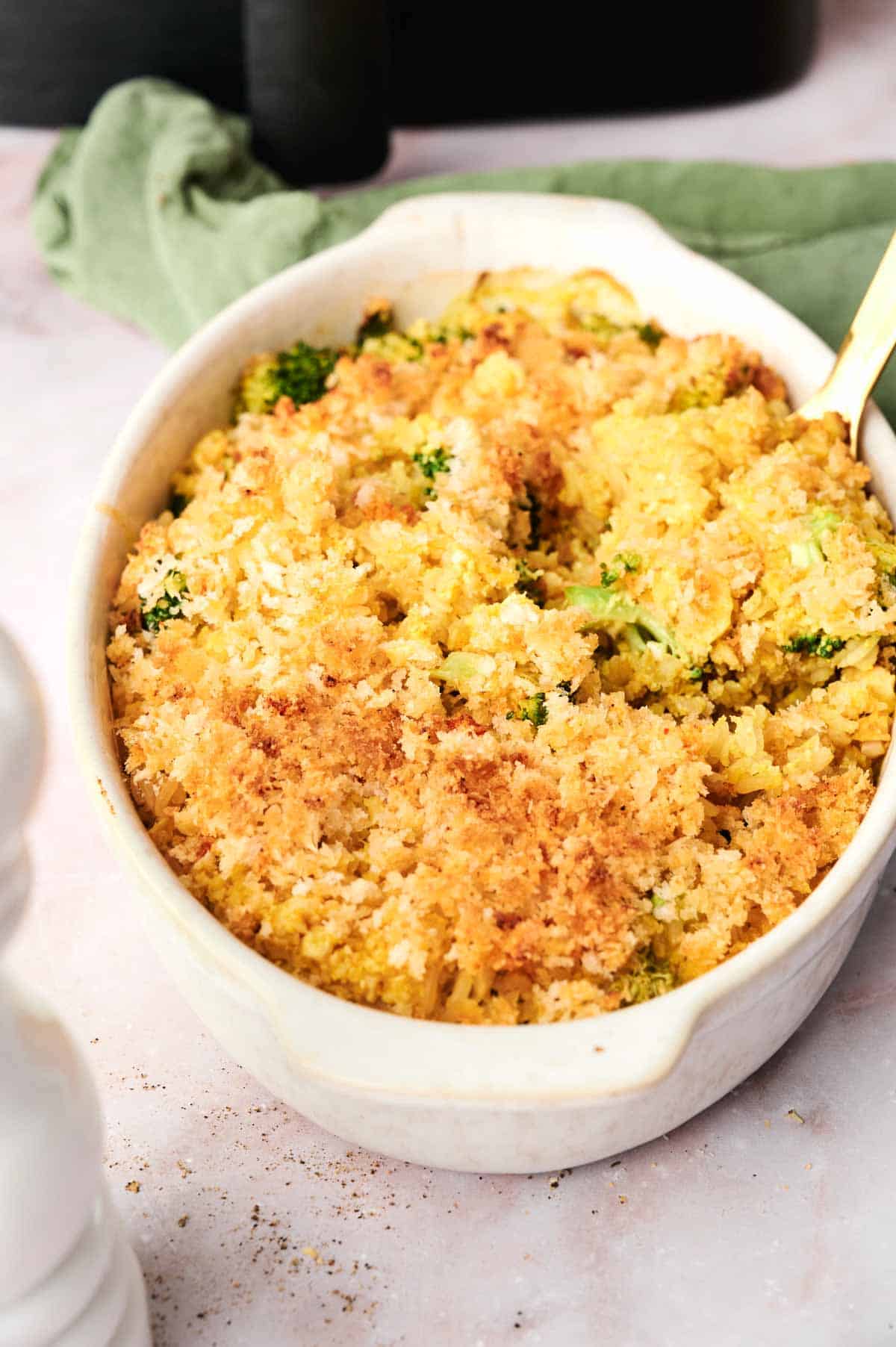 An oval dish holds an Air Fryer vegan broccoli casserole, featuring a creamy rice and cheese-like filling beneath its golden, crispy topping. Nearby, a green cloth and a hint of white add to the cozy kitchen scene.